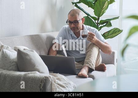 Man working from home, enjoying a cup of coffee while using his laptop on the sofa Stock Photo