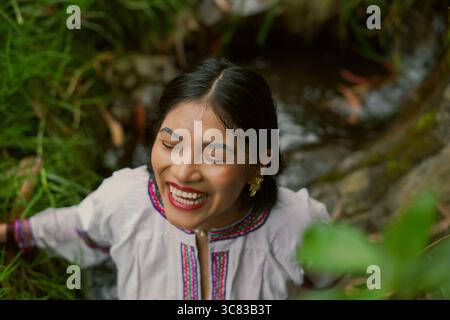 Young indigenous woman laughing joyfully near a river in a lush forest, embracing her culture and connecting with the beauty of nature in a serene, pe Stock Photo