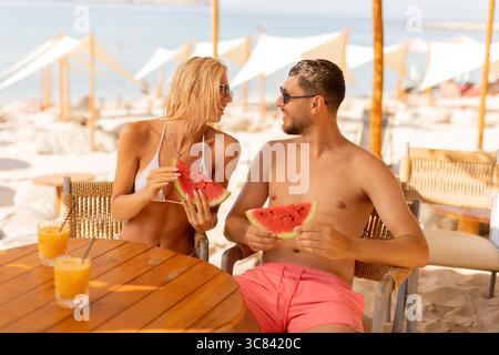 Two people share laughter and fresh watermelon while seated at a beachside table. The sun shines brightly, enhancing their joyful moment amidst tropic Stock Photo