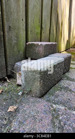 Close-up of stacked concrete bricks resting on a moss-covered outdoor surface, set against a weathered wooden fence tinged with green. Stock Photo