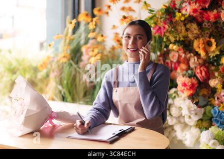Young Asian female florist manages thriving flower shop. She handles customer inquiries via phone while taking important notes. Stock Photo