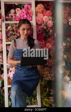 Dedicated Asian adult woman handles digital retail operations in her charming flower boutique focusing on efficiency and growth Stock Photo