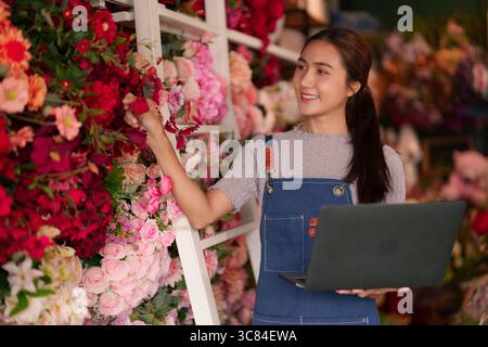 Happy Asian adult woman flower shop owner working digital tasks on laptop managing beautiful floral arrangements vibrant retail store environment Stock Photo