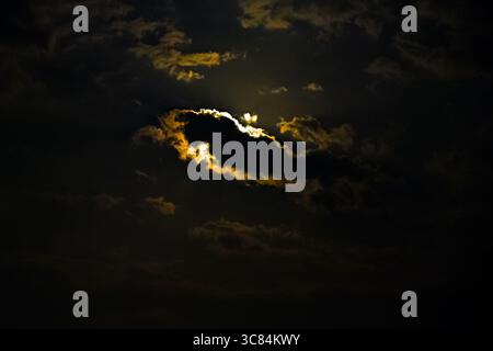 A bright full moon breaks through a dramatic formation of dark storm clouds at night, creating an ethereal glow and moonbeam effect. Stock Photo