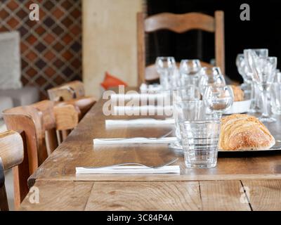 A long rustic wooden table set for a festive indoor meal, with clear glasses, white napkins, and a loaf of sliced artisan bread. Stock Photo