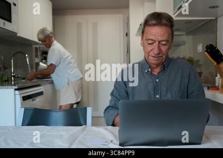 Senior man using laptop while wife washing dishes in kitchen Stock Photo