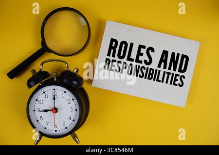 Roles And Responsibilities text written on paper with magnifying glass and alarm clock on yellow background Stock Photo