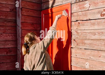Woman applies protective paint to metal door using flat paint brush, ensuring rust resistance for country house entrance. Stock Photo