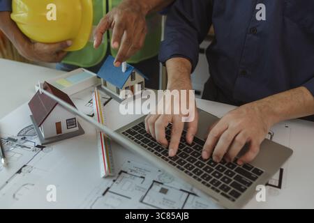 there are scientific calculator, laptop, house model, blueprint on the desk in the office with two architects, engineer is meeting with contractor abo Stock Photo