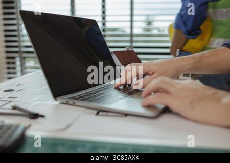 there are scientific calculator, laptop, house model, blueprint on the desk in the office with two architects, engineer is meeting with contractor abo Stock Photo