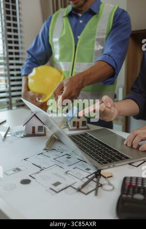 there are scientific calculator, laptop, house model, blueprint on the desk in the office with two architects, engineer is meeting with contractor abo Stock Photo