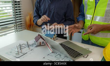 there are scientific calculator, laptop, house model, blueprint on the desk in the office with two architects, engineer is meeting with contractor abo Stock Photo