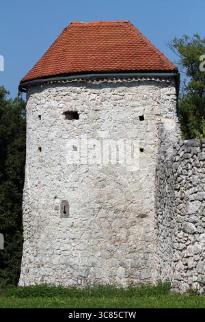 Ancient watchtower built of rough stone with red tile roof, small slit windows for archers, and attached stone wall, surrounded by greenery Stock Photo