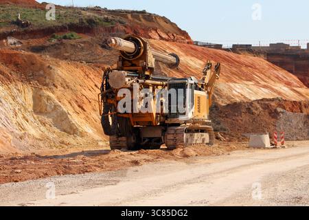 A large rotary drilling rig sits on a dirt road at a multicolored excavation site, displaying tracked wheels, soil-stained metal, and heavy mechanical Stock Photo