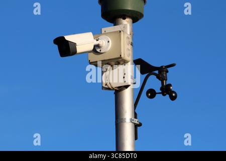 A metal pole supports a white surveillance camera and black anemometer, capturing traffic and weather data, ideal for monitoring road safety in urban Stock Photo