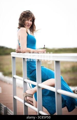 Woman in blue dress leaning on railing holding sunglasses, relaxed summer lifestyle portrait in coastal landscape Stock Photo
