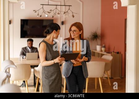 Female colleagues with digital tablet sharing ideas and discussing project details during office meeting Stock Photo