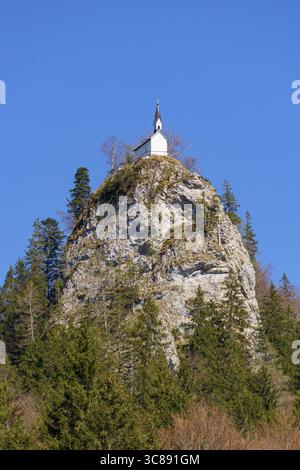 A small white chapel with a black steeple stands atop Riederstein mountain in Bavaria, surrounded by rocky terrain, birch and spruce trees, under a cl Stock Photo