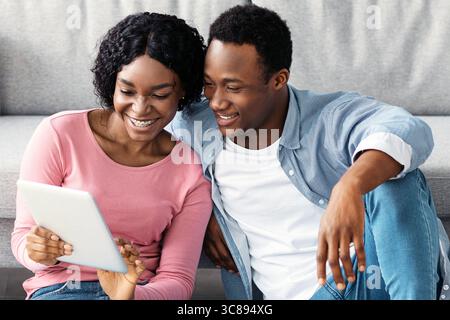 Smiling black couple with cleaning tools resting, using digital tablet Stock Photo