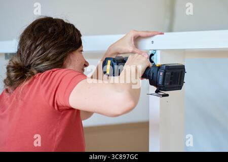 Woman using cordless drill to screw part into white bed frame. Female assembling furniture in new apartment. DIY home renovation and home organization Stock Photo