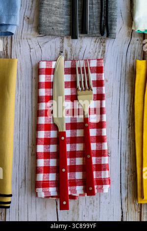 Top view of a table setting with a fork and knife placed on a red and white checkered napkin over rustic wooden background. Colorful folded napkins Stock Photo