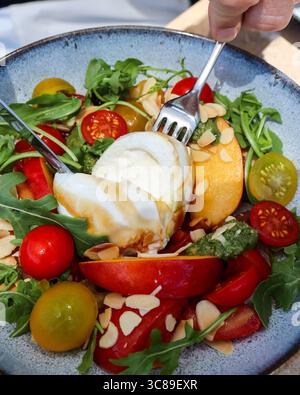 close up of a fresh and colourful burrata salad with cherry tomatoes, peach slices, green leaves and nuts. The burrata is been split open Stock Photo