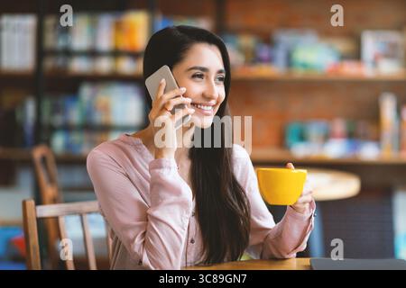 Smiling latin lady talking on mobile phone while resting at cafe alone, looking aside and smiling, copy space Stock Photo