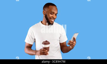 Black Guy Using Phone Holding Paper Coffee Cup, Blue Background Stock Photo