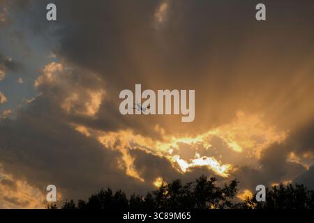 Airplane flying through dramatic sunset clouds with sun rays breaking through Stock Photo