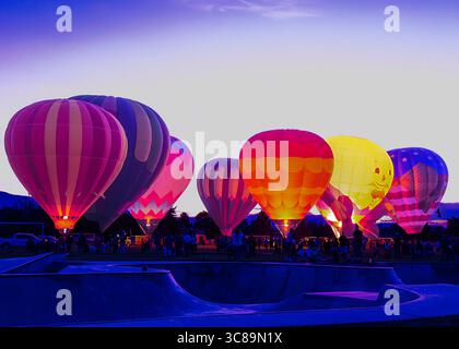 Hot air balloons lit up at night glow in the sky at a balloon festival where people watch from the park below. See glowing designs on the balloons. Stock Photo