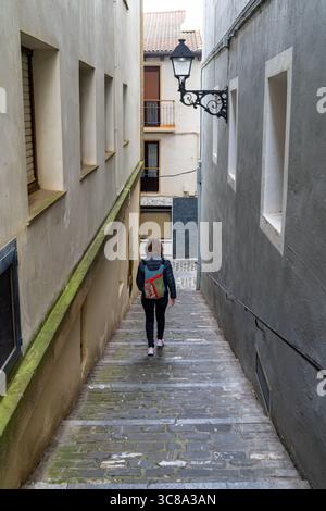 A woman walks down a narrow, stone-paved street between tall buildings with a classic street lamp in a charming European town. Stock Photo