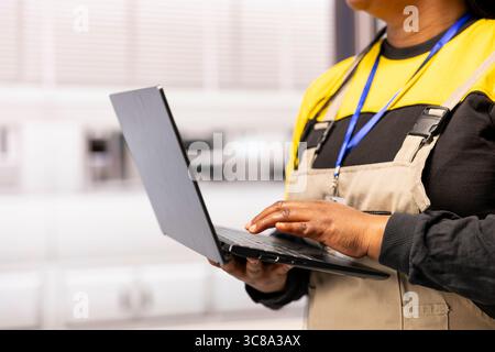 Female engineer enhances efficiency in industrial facility, using laptop to conduct quality control and oversee operation of manufacturing machines. Black technician managing automated production line Stock Photo