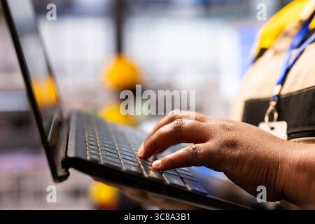 Automation expert operating industry 4.0 software on laptop and ensuring maximum factory efficiency. African american engineer evaluating robotic arms and automated systems in industrial facility. Stock Photo