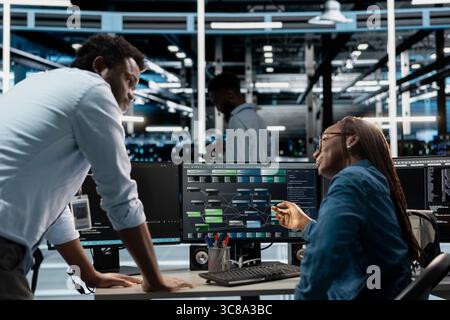 Supervisor checking work done by engineer programming in server hub housing infrastructure storing datasets. African american team leader overseeing IT specialist repairing data center racks Stock Photo