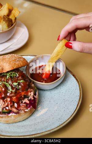 pulled pork sandwich served with french fries and ketchup sauce, a female hand is dipping a potato fry in the sauce Stock Photo