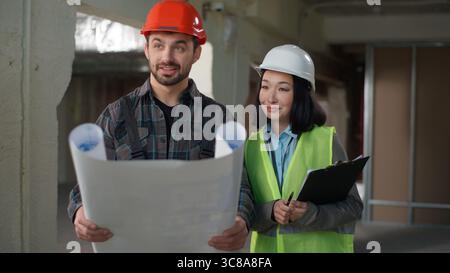Two diverse Caucasian Japanese builders contractors man woman civil engineers architects workers in helmet uniform talking discussing blueprint paper Stock Photo