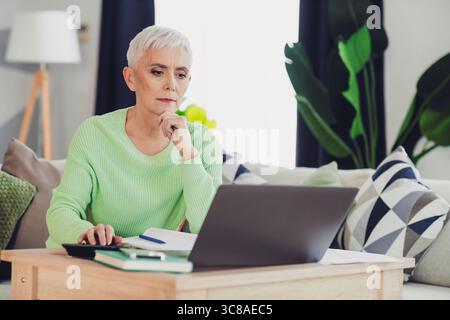 Mature woman using a laptop at home relaxing on a couch. Senior adult working or browsing online in a cozy living room. Stock Photo