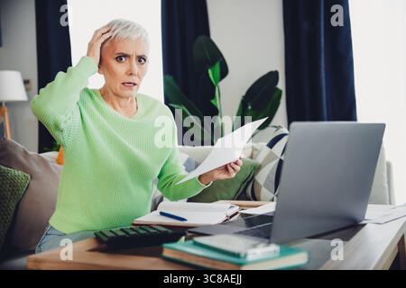 Mature woman in bright sweater using laptop and reading documents while sitting on sofa in modern living room Stock Photo