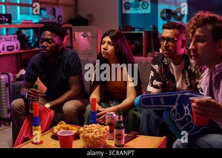Group of young adults including Black man, Asian woman, Caucasian men sitting on couch watching television, holding drinks and snacks, engaging in social gathering, focused on screen Stock Photo
