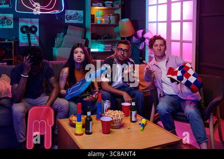 Group of four young adults, including Black man, Asian woman, South Asian man, and Caucasian man, sitting on couch watching television, holding snacks and drinks, showing mixed emotions Stock Photo