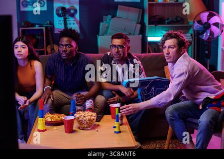 Group of young adults including Caucasian man, Black man, South Asian man, and Hispanic woman watching television together, holding snacks and drinks, sitting around table Stock Photo