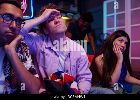 Group of young adults and one Black man in background watching screen with bored expressions, multiethnic friends sitting together indoors, showing disappointment or frustration Stock Photo