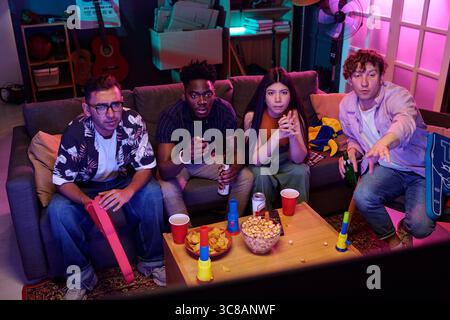 Group of young adults including Caucasian man, Black man, Asian woman, and another Caucasian man watching television together, holding snacks and drinks, sitting on couch in living room Stock Photo