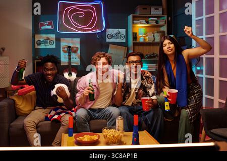 Group of young adults including Black man, Caucasian man, South Asian man, Latina woman sitting on couch watching sports game, cheering, holding drinks, celebrating together indoors Stock Photo