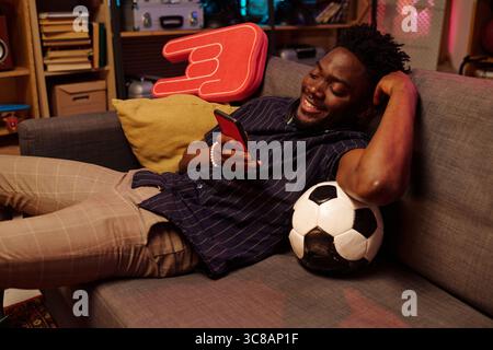 Young adult Black man reclining on sofa holding smartphone and smiling, resting arm on cushion with soccer ball by side, appearing relaxed in casual home setting Stock Photo