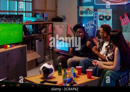 Young adult Black man holding laptop and celebrating with young adult South Asian man and young adult Caucasian woman Stock Photo