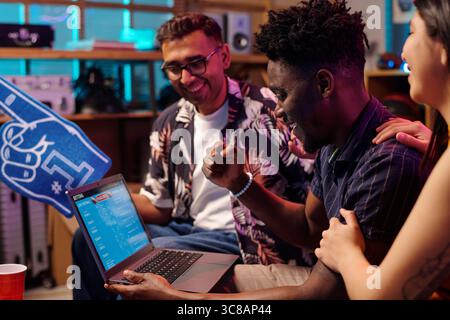Group of young adults including Black man, Caucasian man, and Asian woman celebrating while watching sports betting results on laptop, smiling and interacting in casual setting Stock Photo