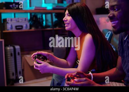 Young adult Caucasian woman and young adult Black man playing video game together, both holding controllers and smiling, sitting side by side in modern indoor setting Stock Photo