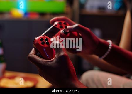 Black young adult man holding video game controller with both hands, playing console game indoors, close up on hands and device, focus on gaming activity, blurred background visible Stock Photo