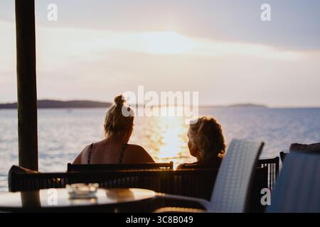 Silhouettes of two unrecognizable middle-aged ladies chatting while sitting on chairs near sea beach at  sunset. Concept of senior leisure. Friends on Stock Photo